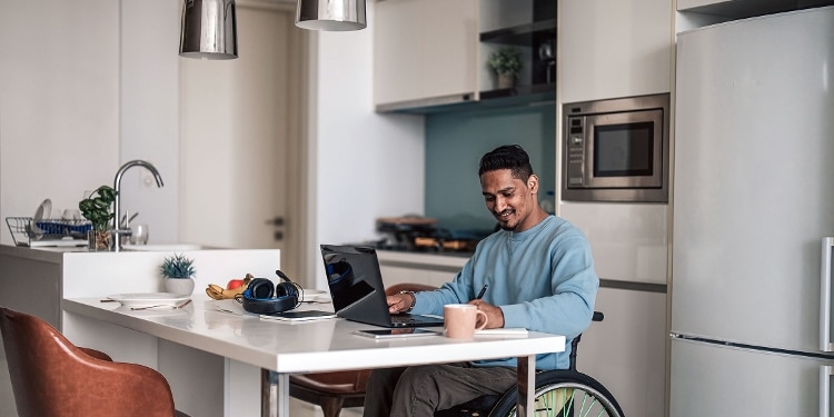 A smiling person sitting in a wheelchair in their kitchen and writing with a cup of coffee and their laptop in front of them.