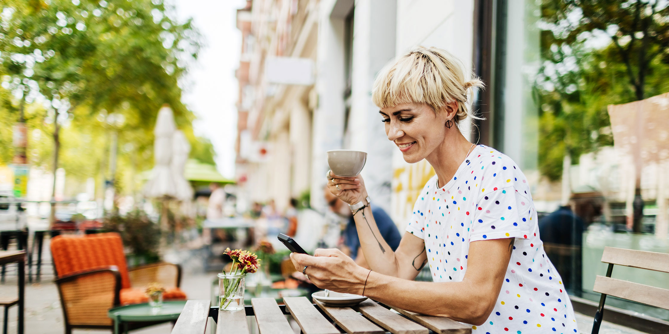 A person sits at a table outside looking at a cellphone with a mug in the other hand.