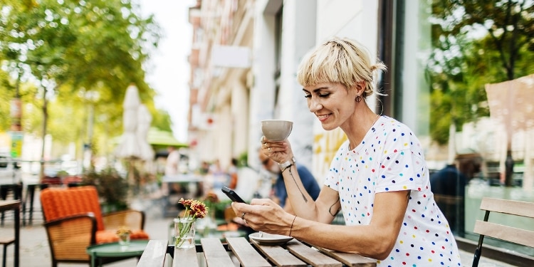 A person sits at a table outside looking at a cellphone with a mug in the other hand.