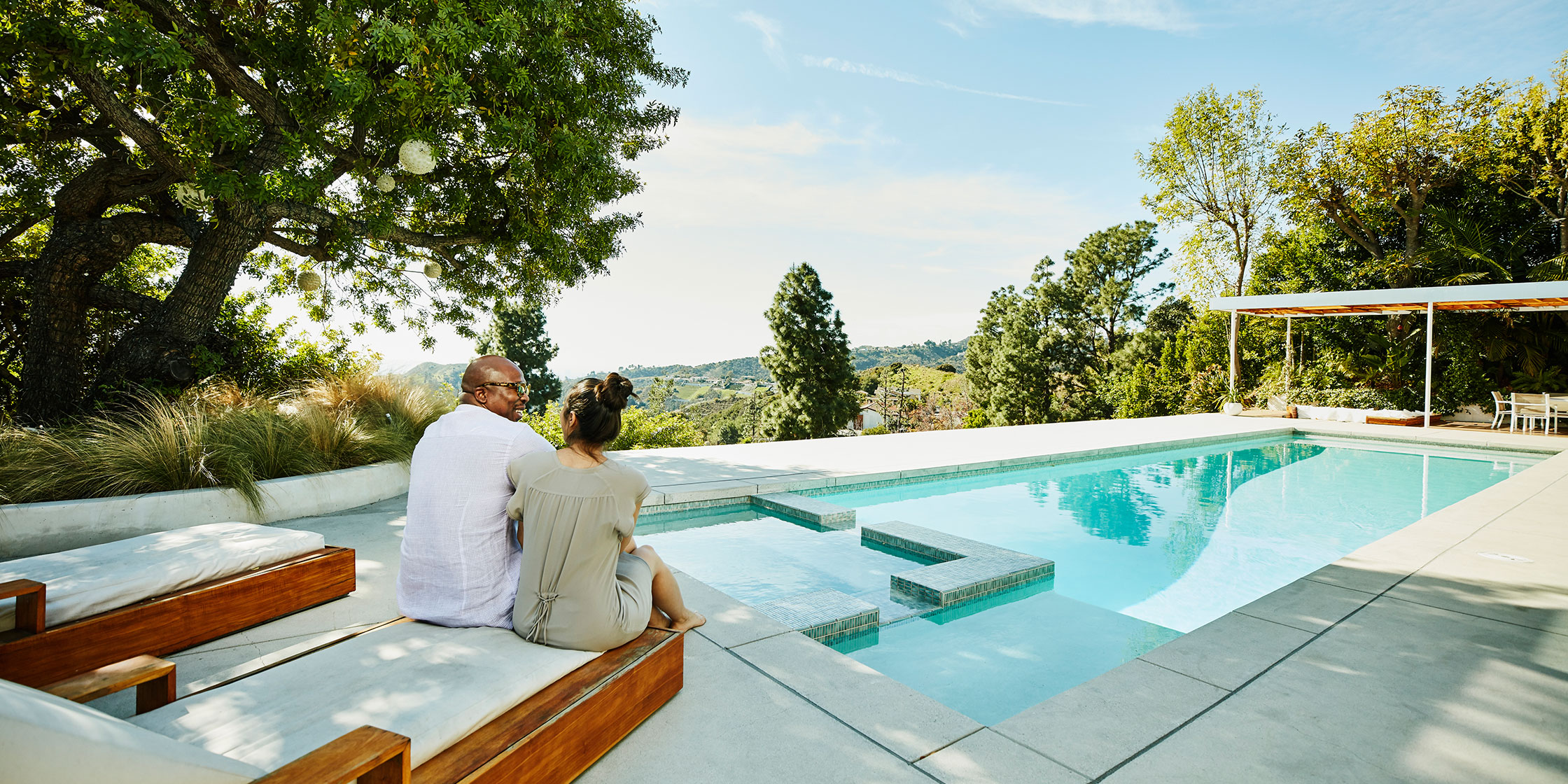 A couple sitting together on the end of a lounger beside a hilltop pool.