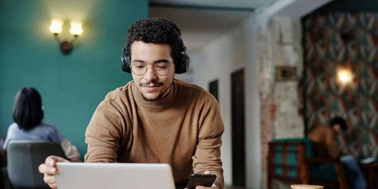 Student wearing headphones holds a cellphone and looks at a laptop.