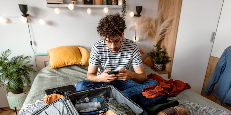 A photo of a person sitting on a bed, checking their phone and packing a suitcase.