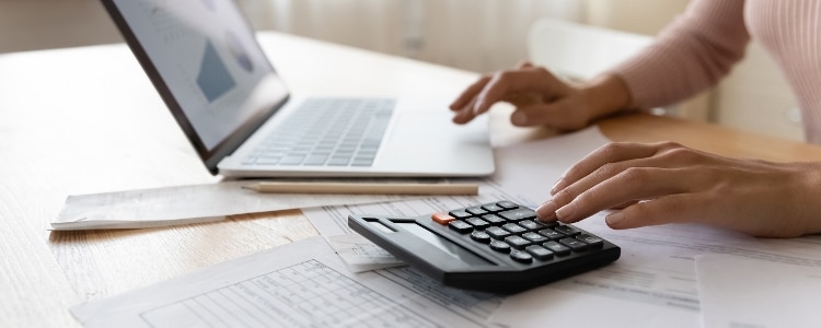 A person sitting at a table calculates their net income using a laptop and calculator.