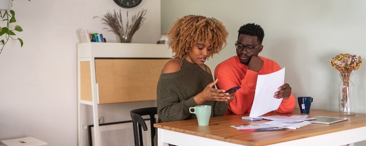 Two people sitting at a table looking at a piece of paper and a cellphone, talking about net worth and how to calculate it.