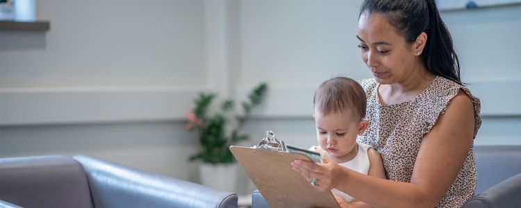 A parent with a baby on her lap sits at a doctor’s office and fills out a form with personal information.