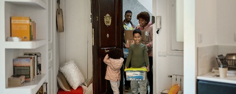 A family of four walks into their new rental home while carrying boxes.