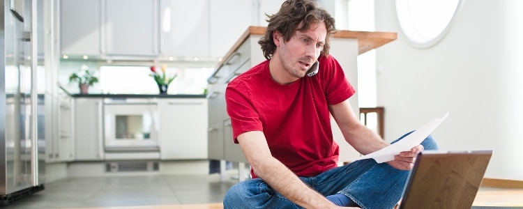 A person sits on the floor while on the phone and looking at a laptop for information about bankruptcy.