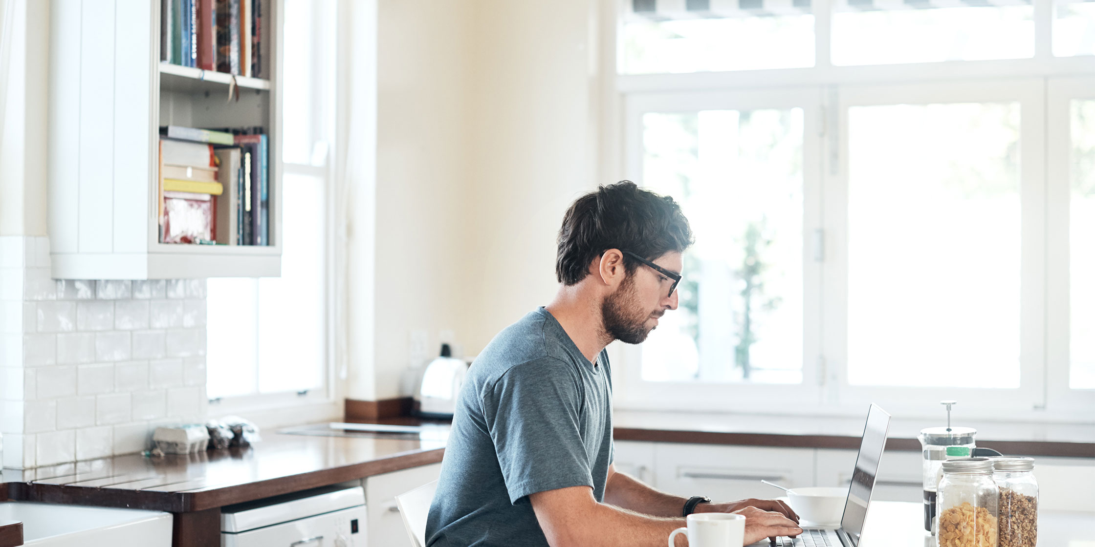 A person in glasses working on their laptop while sitting in their kitchen.