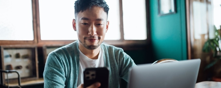 Smiling person sitting with their laptop and looking at their cellphone.