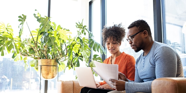 Two people sit on a couch while using a laptop and looking at a stack of documents.