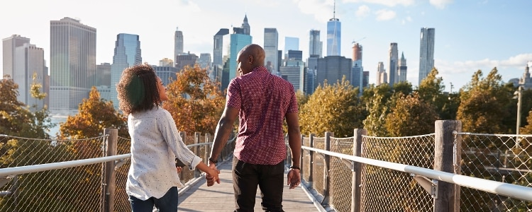 A couple holds hands while walking on a footbridge toward a city skyline.