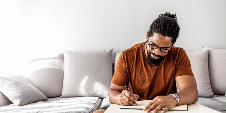 A man wearing glasses and a watch sits on his couch and writes.
