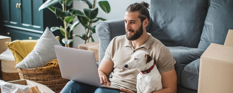 Man sits on the floor against his sofa, his dog beside him and laptop on his knees, to start notifying organizations of his address change.