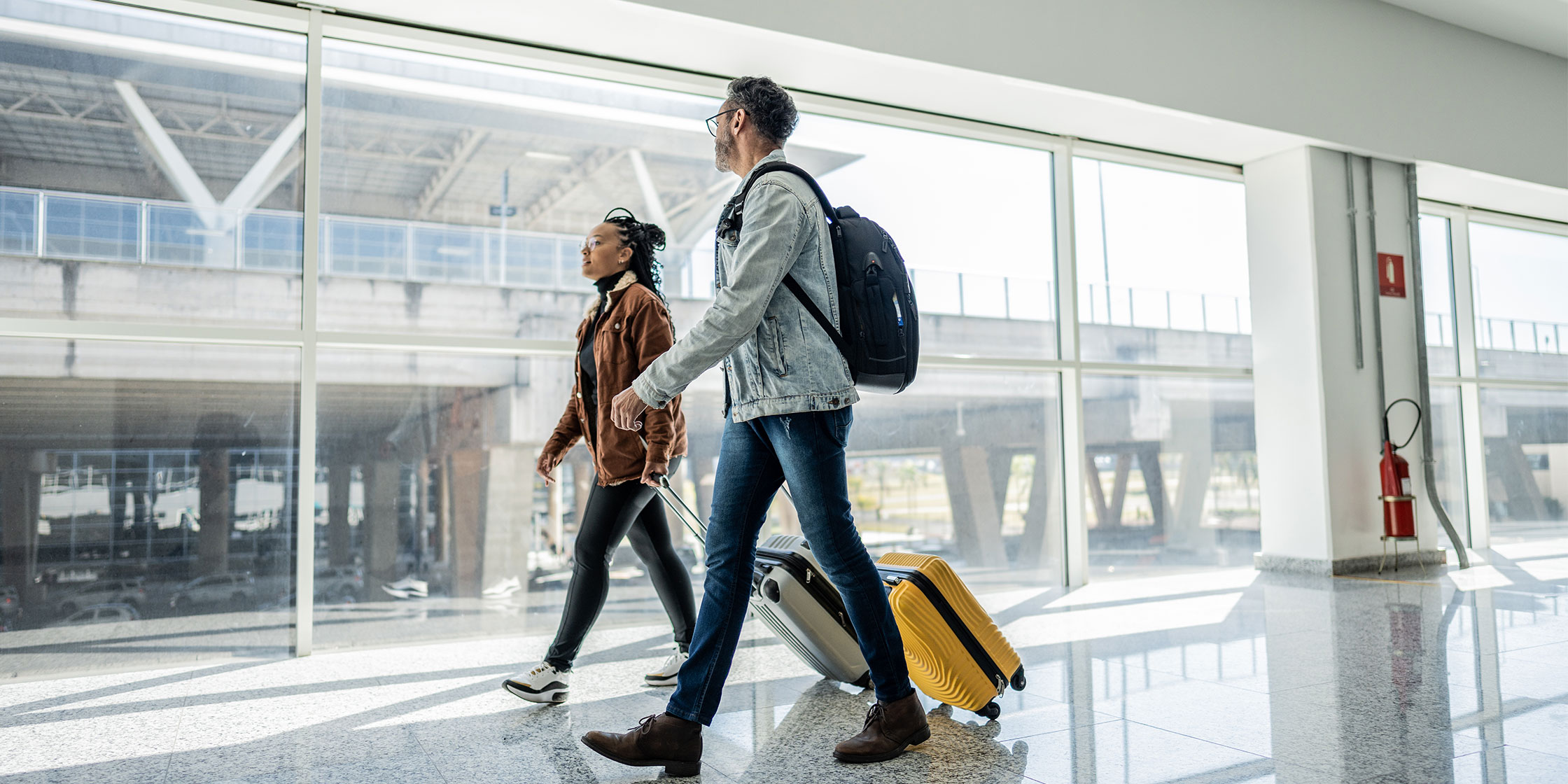 Two people walk through a sunny airport terminal on the way to the security checkpoint.