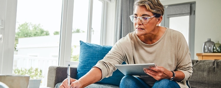 A person sitting on a couch while holding a tablet and taking notes with a pen and paper.