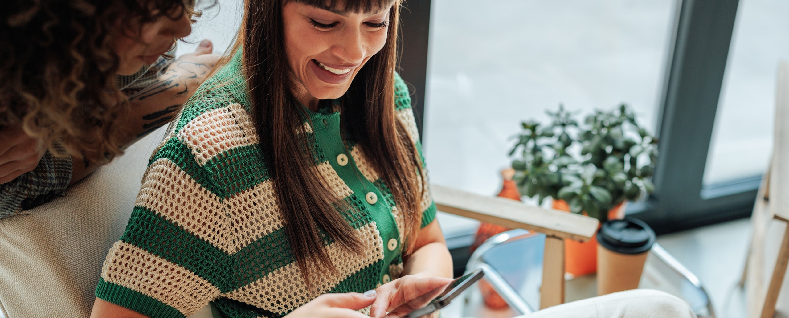 A smiling woman looks at her smartphone while another person leans in to see the screen in a bright room.