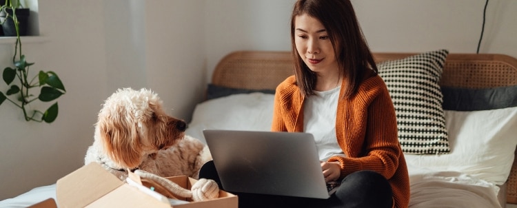A woman in an orange cardigan sits on a bed using a laptop next to her dog and an open delivery box.