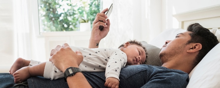 Adult lying on a bed while looking at phone with infant sleeping on top of him.