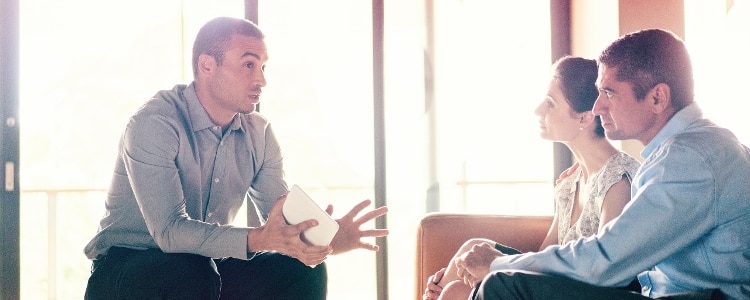 Person holding a tablet while talking to two people seated on a couch.