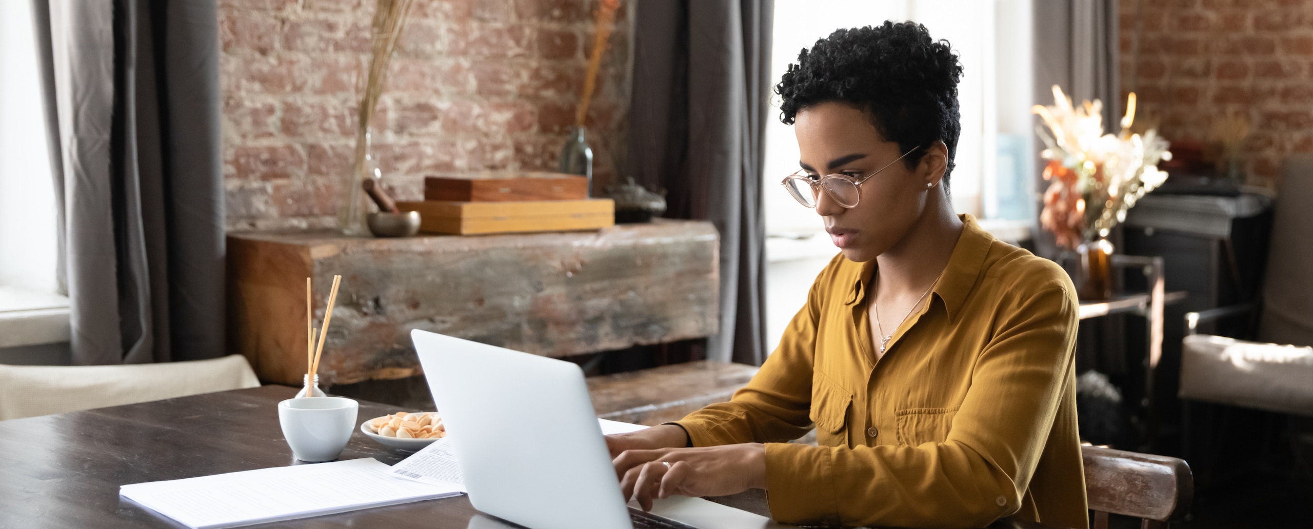 A woman in a yellow long-sleeve shirt sits at a desk while typing on her laptop.