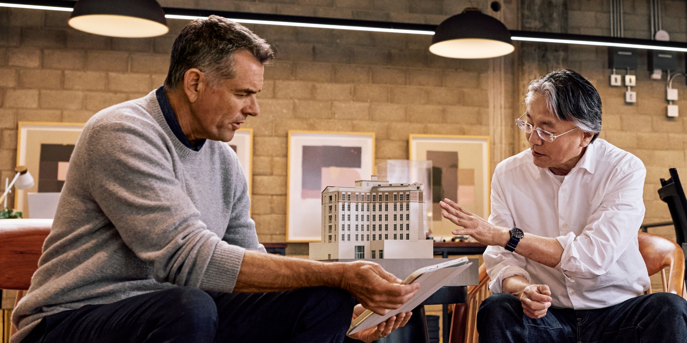 Two men sit together. One is holding out a digital tablet that they’re both looking at. There’s a model of a tall apartment building on the table  behind them.