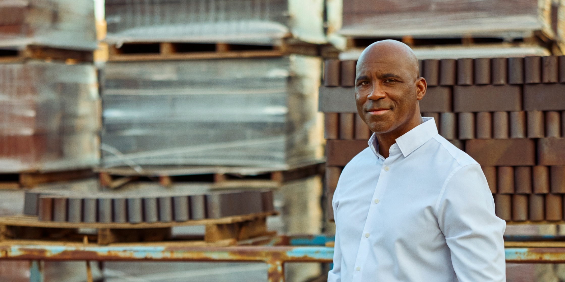 A man in a white button-down shirt stands in front of pallets.