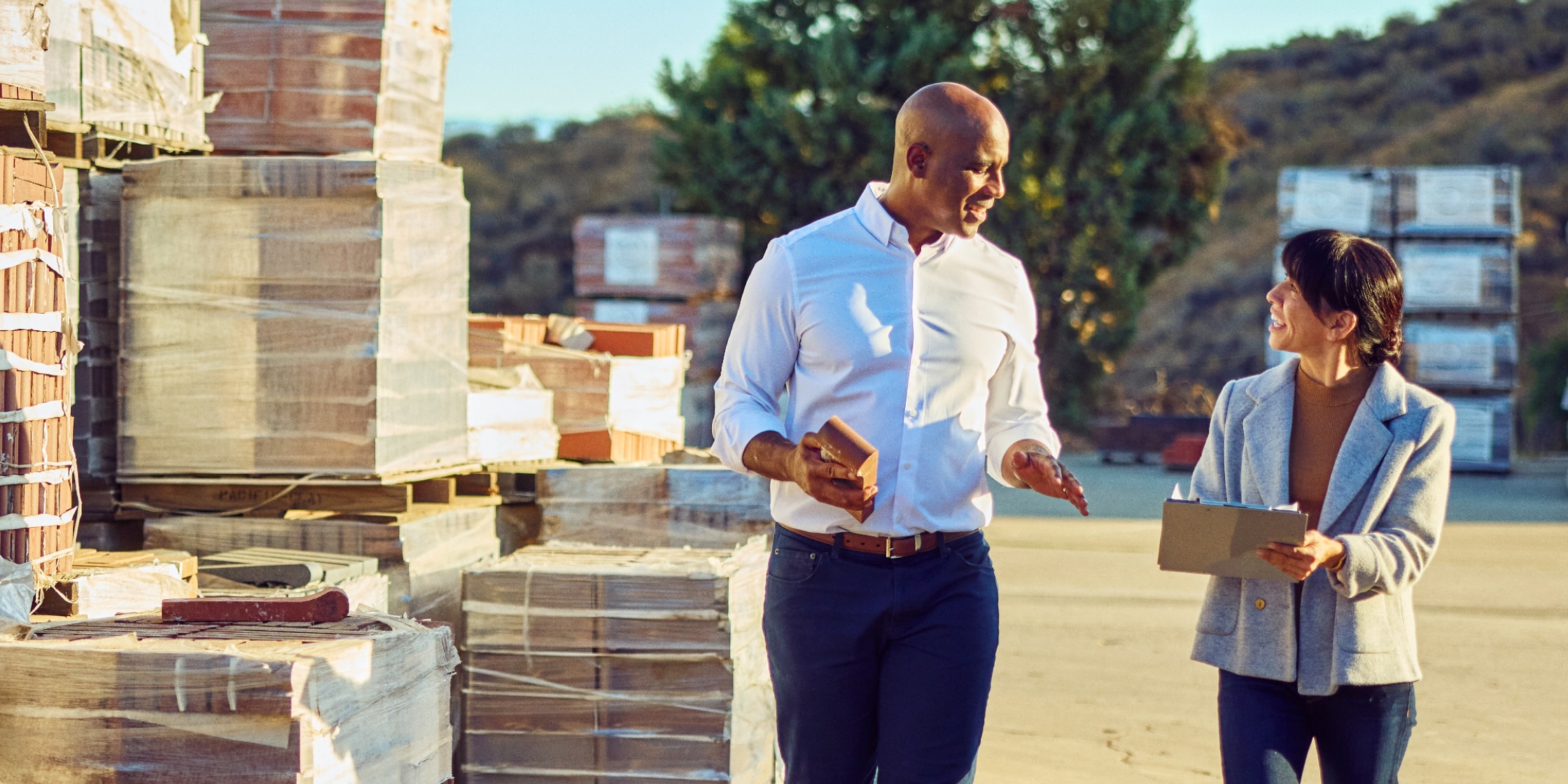 A man and woman walk through a construction site together. There are stacked pallets beside them, and the woman is holding a clipboard.