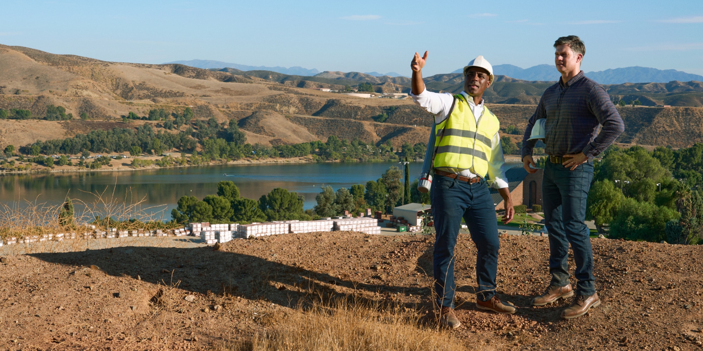 Two men stand together. One is wearing a hard hat and yellow construction vest. The other is in jeans and a plaid shirt. There is a lake and hills behind them.