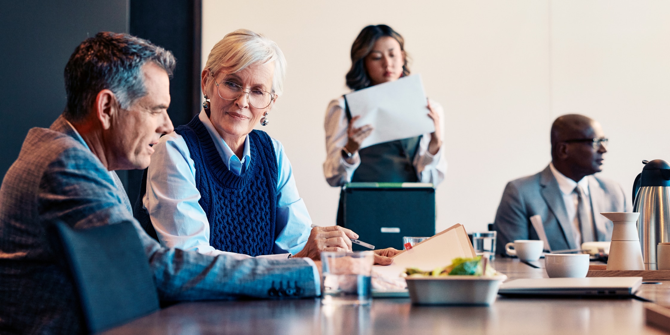 A group of people sit around a conference room table. A woman sifting through papers stands in the background.