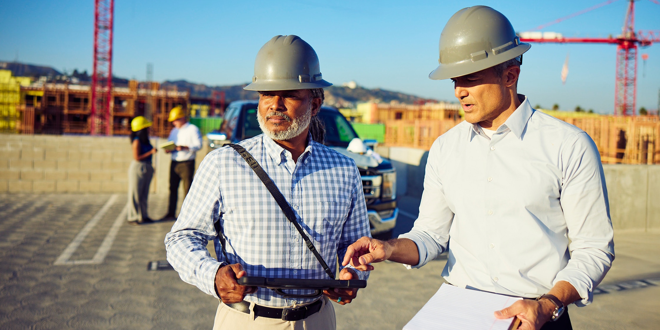 Two men stand together in a construction site. They’re both wearing hard hats and looking at a digital tablet.