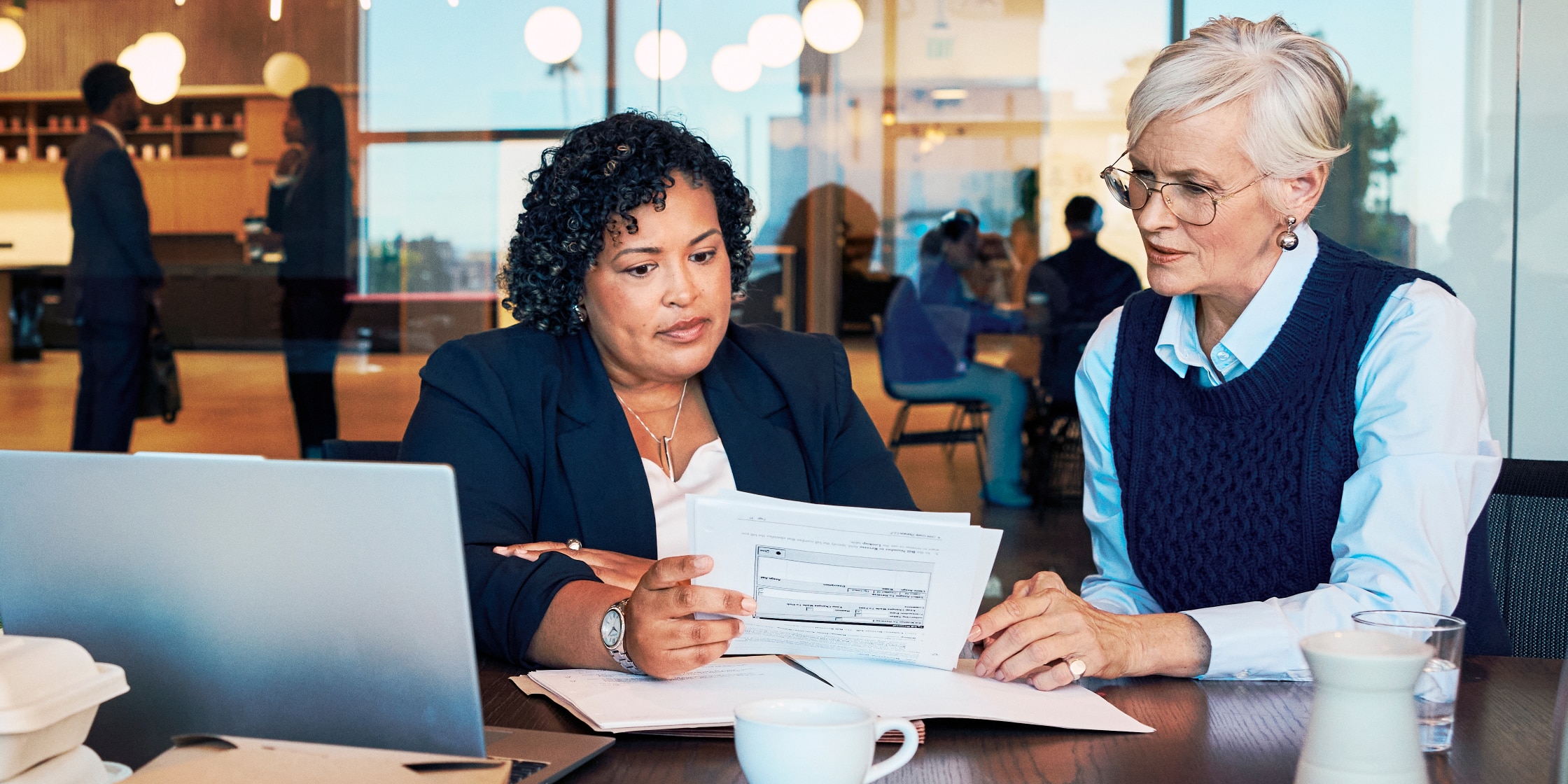 Two women look over paperwork while sitting at an office table together.