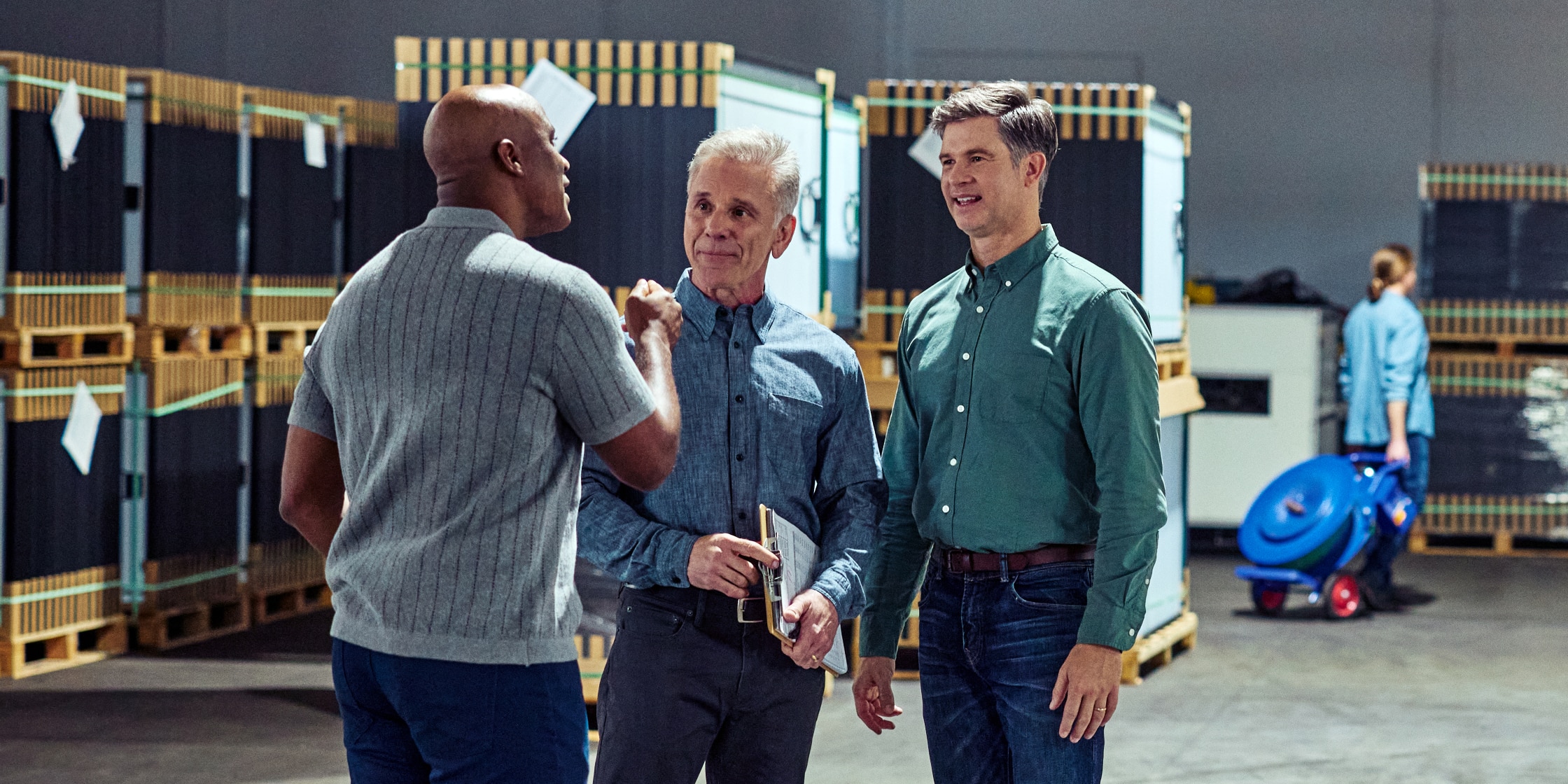 Three men stand together in a warehouse. There are large pallets behind them and a worker in the background.