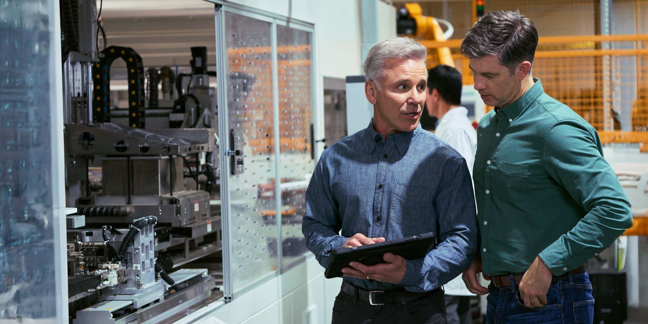 Two men stand in a laboratory together. They’re wearing button-down shirts, and someone in a lab coat is in the background.
