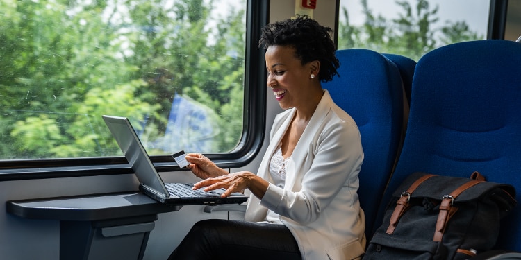 A woman is sitting on a train with her laptop open on the tray table in front of her. She has her credit card out.