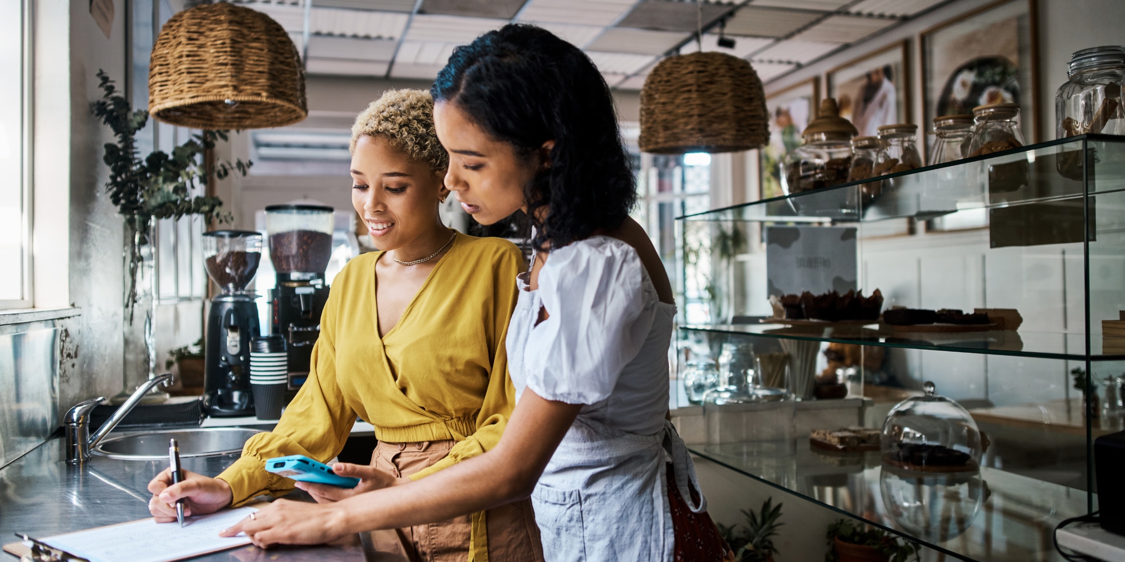 Two women stand in a shop together looking at an iPhone and comparing it to a spreadsheet.
