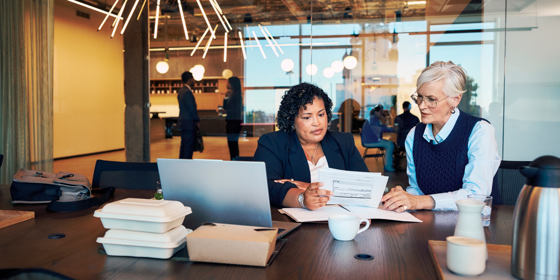 Two women sit at an office conference table with an open laptop in front of them while looking through documents together.