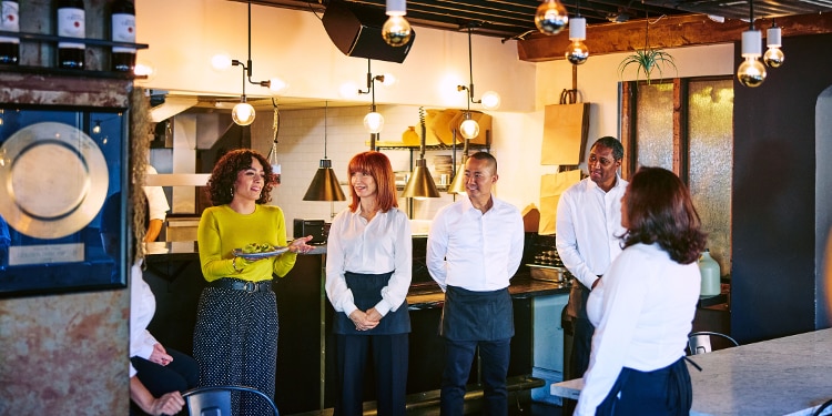 Business owner having a standing meeting with restaurant serveres in front of the kitchen.