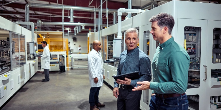 Two men talk while walking together in a laboratory. There are people in white lab coats in the background.