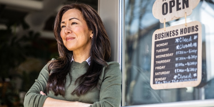 A woman in a green sweater stands next to the OPEN sign of her business.
