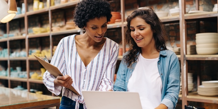 Two women who work in a pottery studio look over a laptop together. The woman on the left is holding a clipboard.