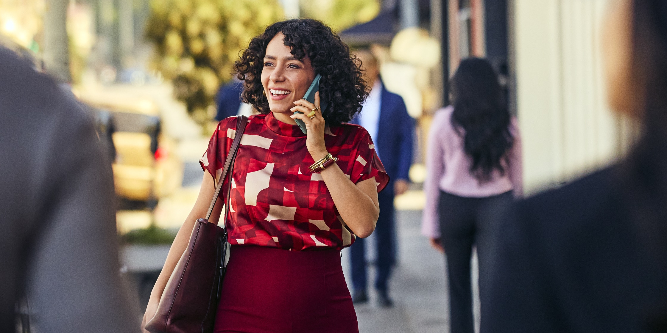 A woman in a red blouse and red pencil skirt walks down the street while talking into her cellphone.