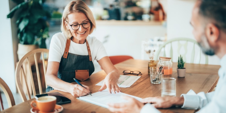 A woman in an apron sits at a table in a cafe. She’s signing paperwork. There’s a man out of focus on the other side of the table.