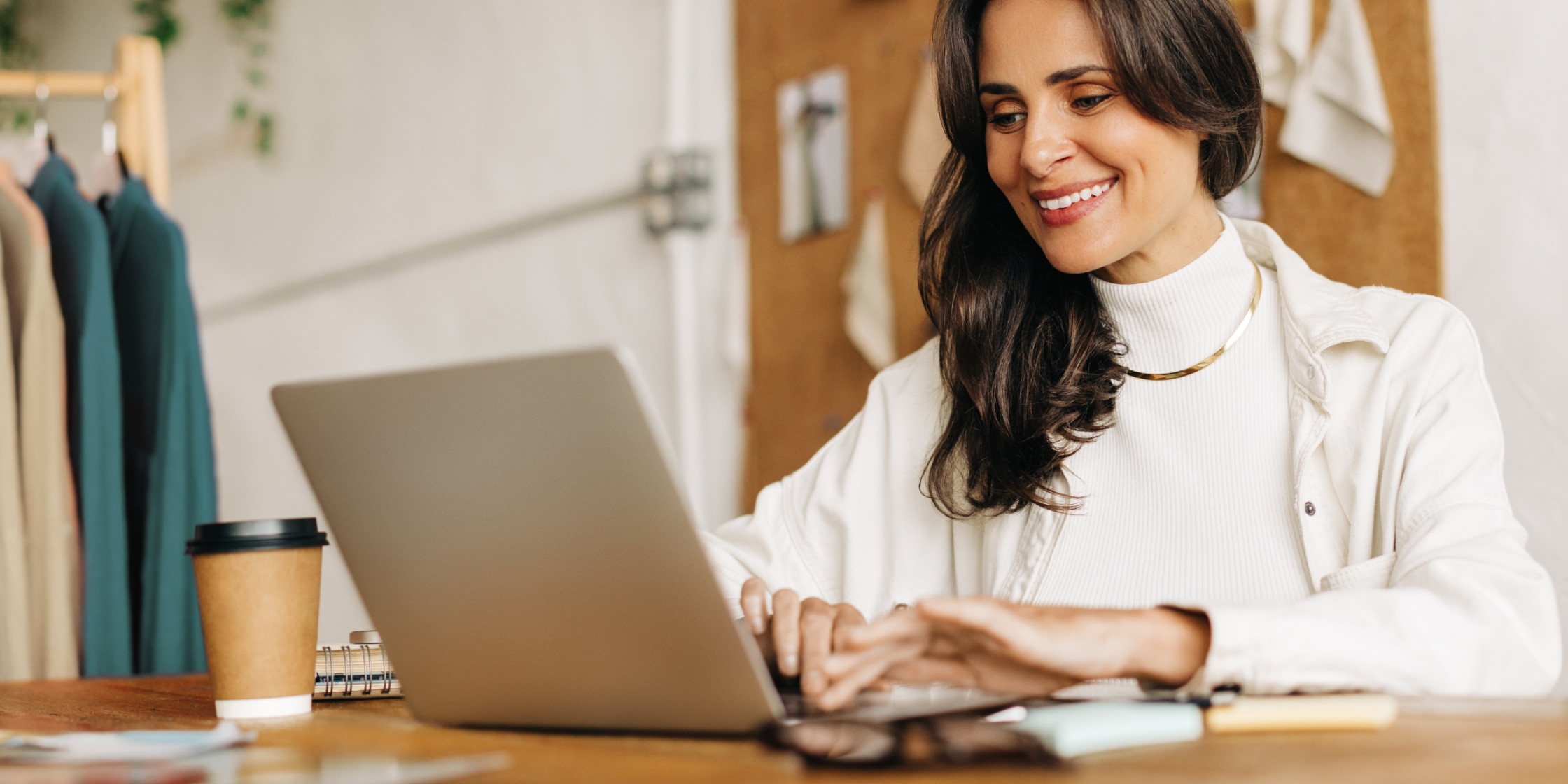 A woman in a white sweater and jacket works on a laptop sitting at a desk.