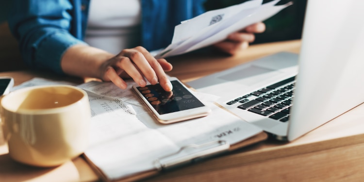 A desk is filled with an open laptop, a coffee mug, paperwork, and a mobile phone open to a calculator, with someone in the background typing in numbers.