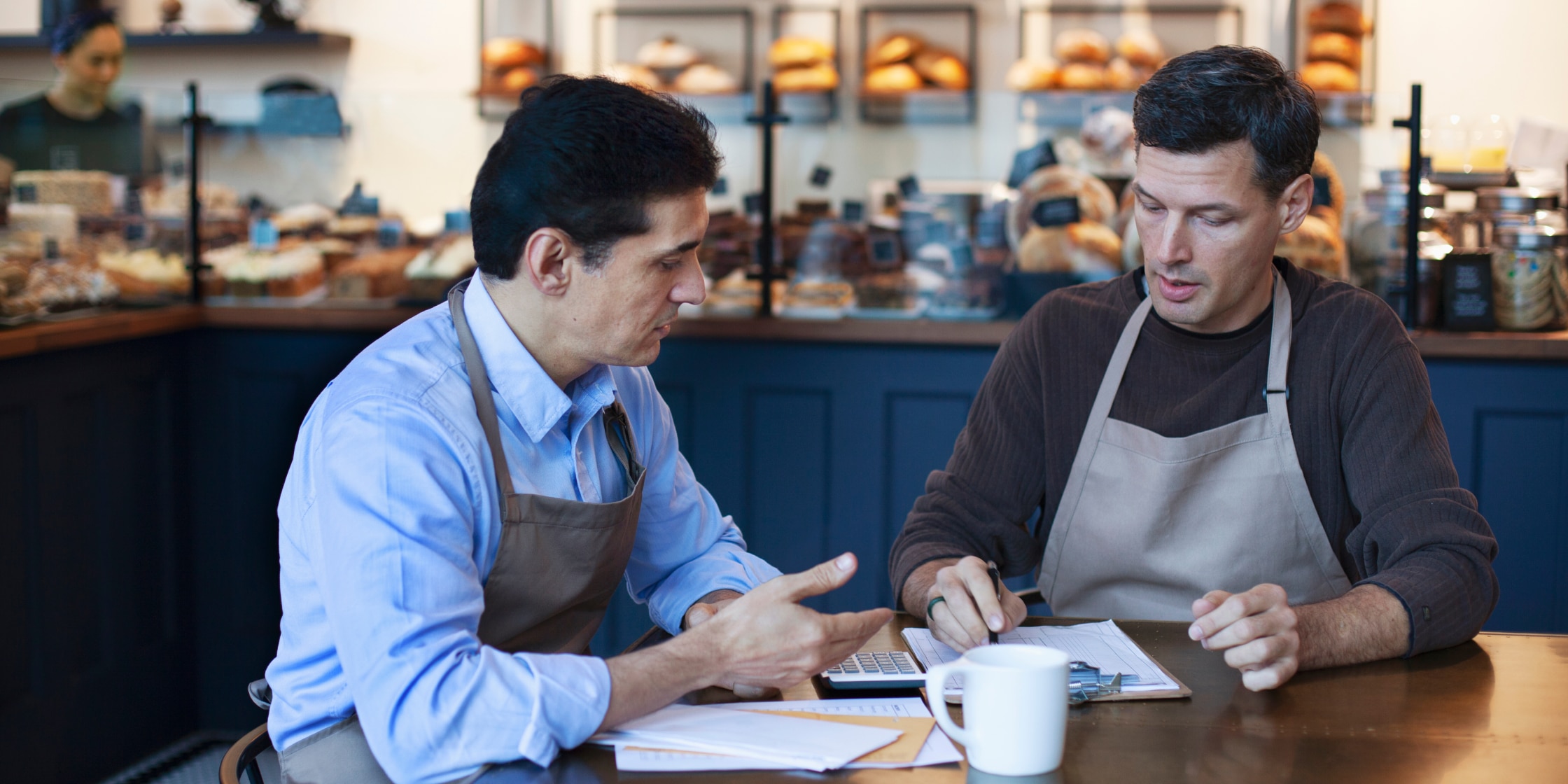Two men sit at a table in a bakery. They’re both wearing aprons and looking over paperwork together.