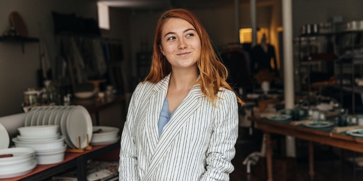 A woman in a white blazer with red hair stands in a studio smiling.