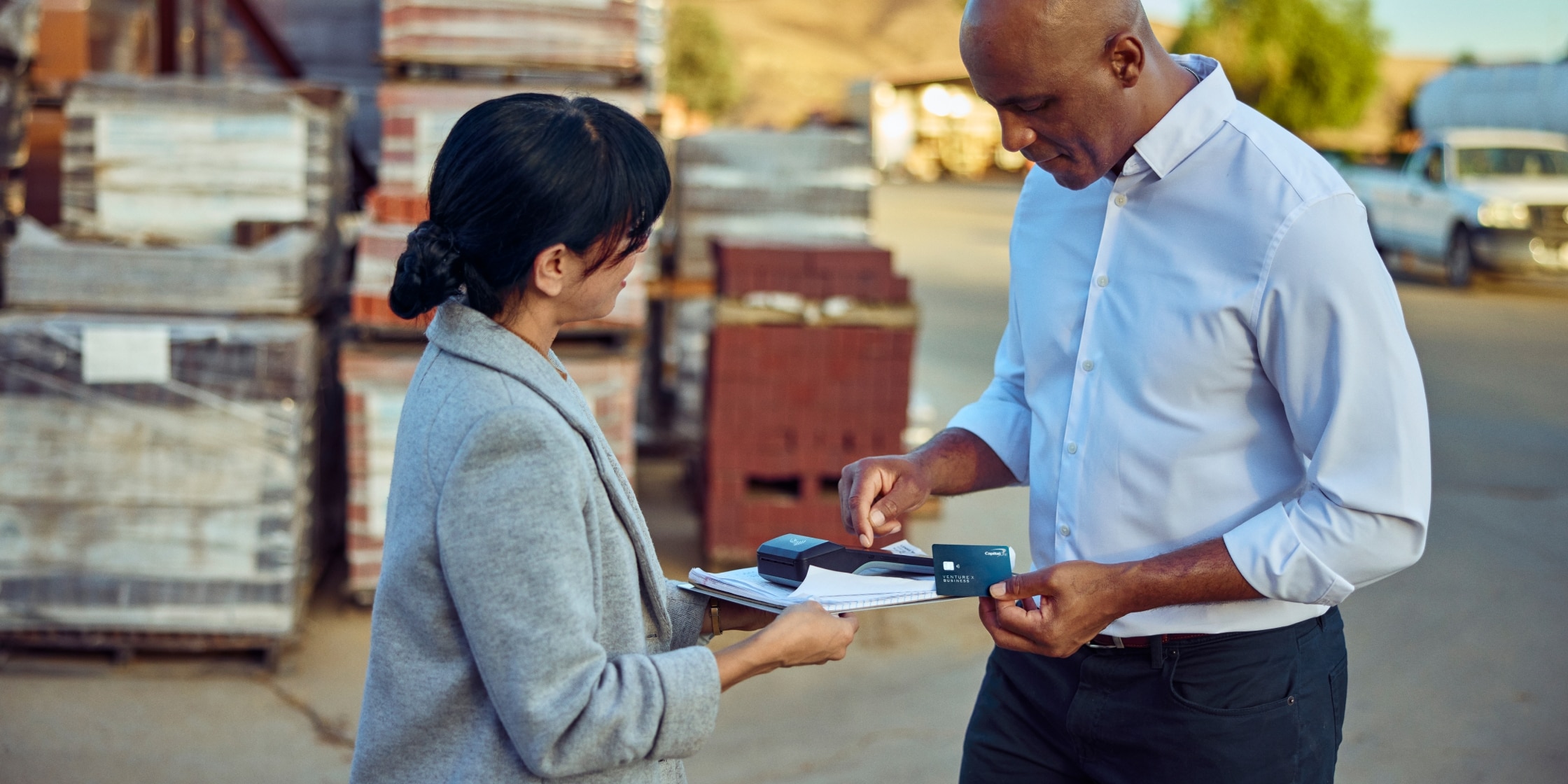 A woman in a gray blazer holds out a credit card reader to a man. The man is holding a blue Capital One Venture X Business credit card.