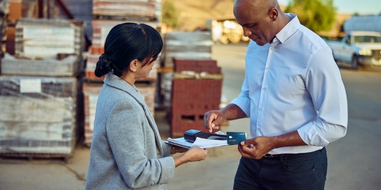 A woman in a gray blazer holds out a credit card reader to a man. The man is holding a blue Capital One Venture X Business credit card.