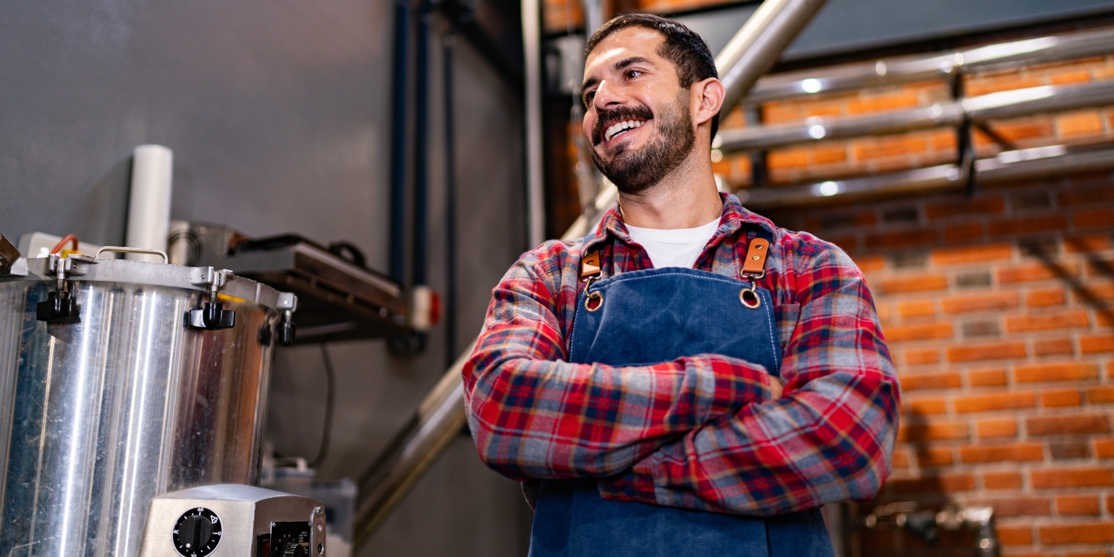 A man stands in an industrial kitchen. He’s wearing a red flannel shirt and a denim apron.