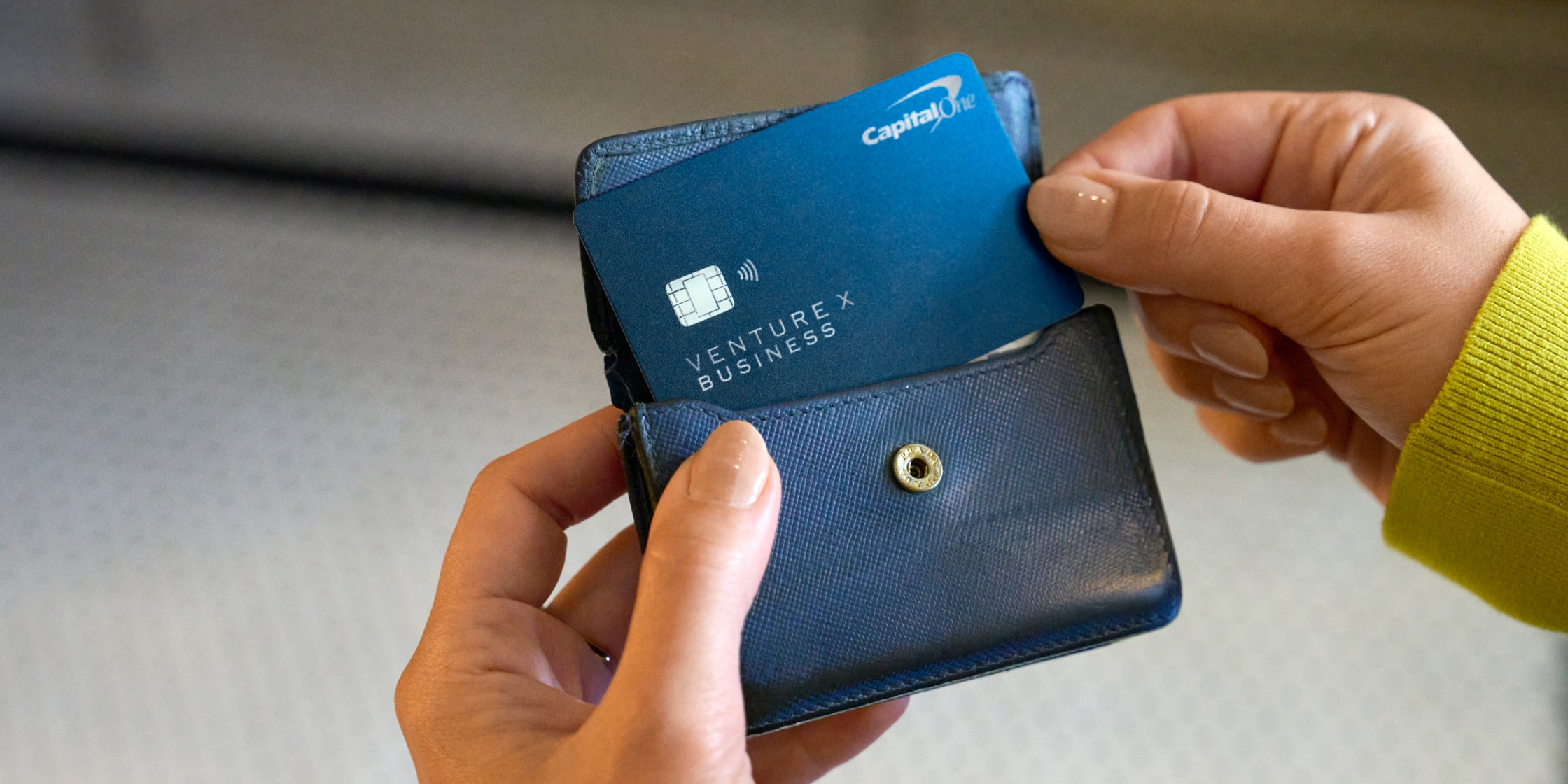 A close-up of a hand holding a small wallet and pulling out a blue Capital One Venture X Business card.